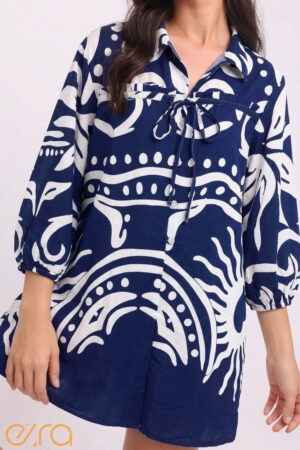 Model wearing an Ezra brand mini tunic dress featuring a bold abstract sunburst and swirling line pattern in deep umber brown on an ivory background. The dress has a button-down front, classic collar, and three-quarter length sleeves with elastic cuffs.