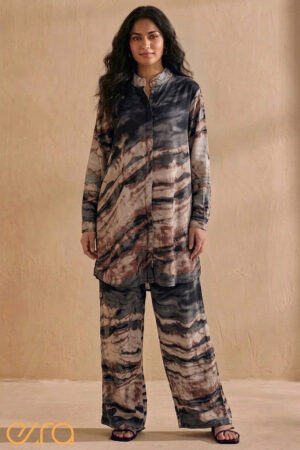 A woman posing in a charcoal and earthy-toned Ezra co-ord set featuring a marble-like abstract landscape print. The ensemble includes a long-sleeved, button-down tunic with a mandarin collar and matching wide-leg fluid trousers, set against a neutral, warm-toned textured wall.