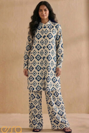A woman in an Ezra brand co-ord set featuring a navy blue and cream geometric diamond ikat print. The outfit includes a long button-up tunic with a mandarin collar and matching wide-leg trousers, standing against a neutral sand-colored background.