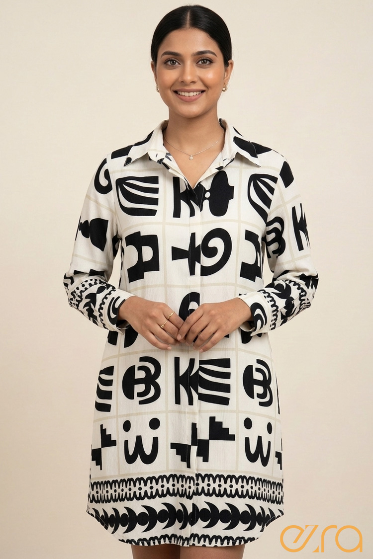 A woman modeling a mini shirt dress featuring a bold monochrome abstract graphic print. The dress is ivory with black geometric and symbolic motifs arranged in a grid pattern, featuring a classic collar, long sleeves with ornate cuffs, and a decorative border hem.
