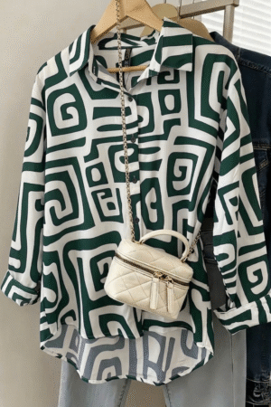 A navy blue and ivory oversized shirt dress with a bold, interlocking spiral geometric print hanging on a wooden hanger. The dress features a classic collar and button-down front, styled with a small cream quilted crossbody bag draped over it