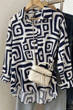 A navy blue and ivory oversized shirt dress with a bold, interlocking spiral geometric print hanging on a wooden hanger. The dress features a classic collar and button-down front, styled with a small cream quilted crossbody bag draped over it