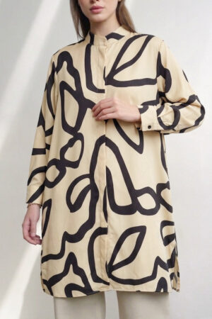 A woman modeling a black, long-sleeved satin tunic dress featuring a bold, abstract mustard-gold swirl pattern, paired with light beige trousers against a neutral background.