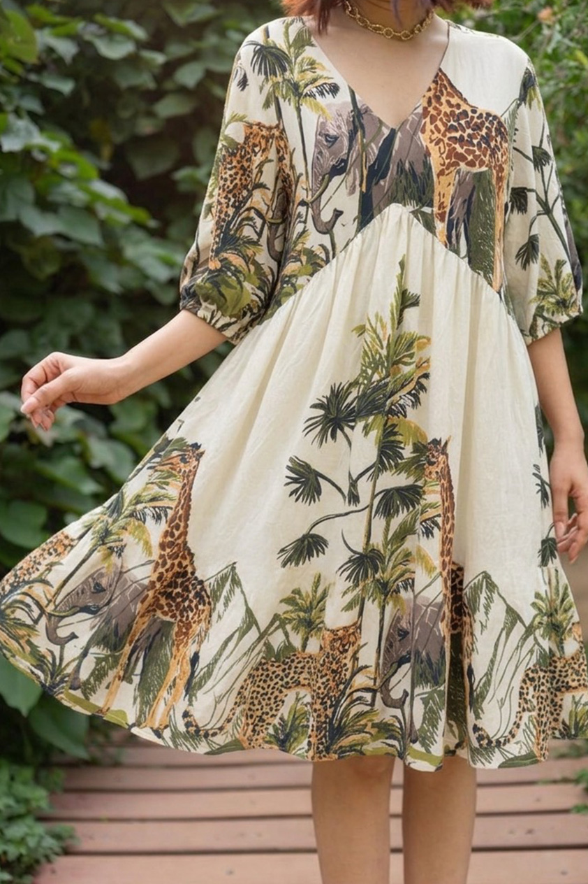 woman models a knee-length cream cotton dress featuring an empire waist silhouette and an illustrated wildlife safari print with giraffes, elephants, cheetahs, and palm trees, styled with a deep V-neckline and three-quarter sleeves while standing on a wooden garden walkway.