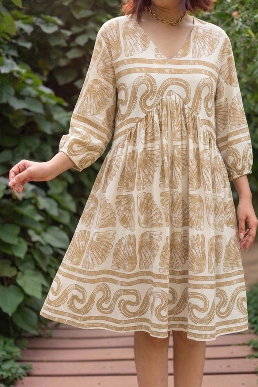 a woman models a knee-length cream cotton dress with an empire waist silhouette, decorated in a repeating ochre-brown seashell and Greek wave block print, designed with a V-neckline and elasticized three-quarter sleeves while standing on a outdoor wooden walkway.