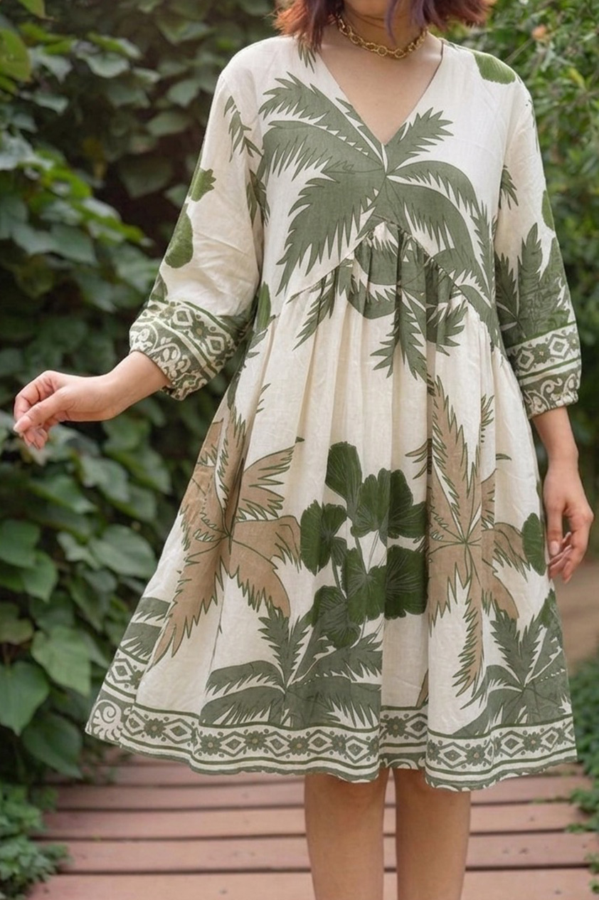 A woman models a knee-length cream cotton dress featuring an empire waist silhouette and a bold olive green and tan tropical palm leaf print, styled with a V-neckline, three-quarter sleeves with geometric borders, standing on a wooden garden walkway.