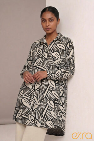A woman leaning against a wall wearing a long sleeve Ezra tunic shirt with a black and cream abstract hand-drawn geometric batik pattern,