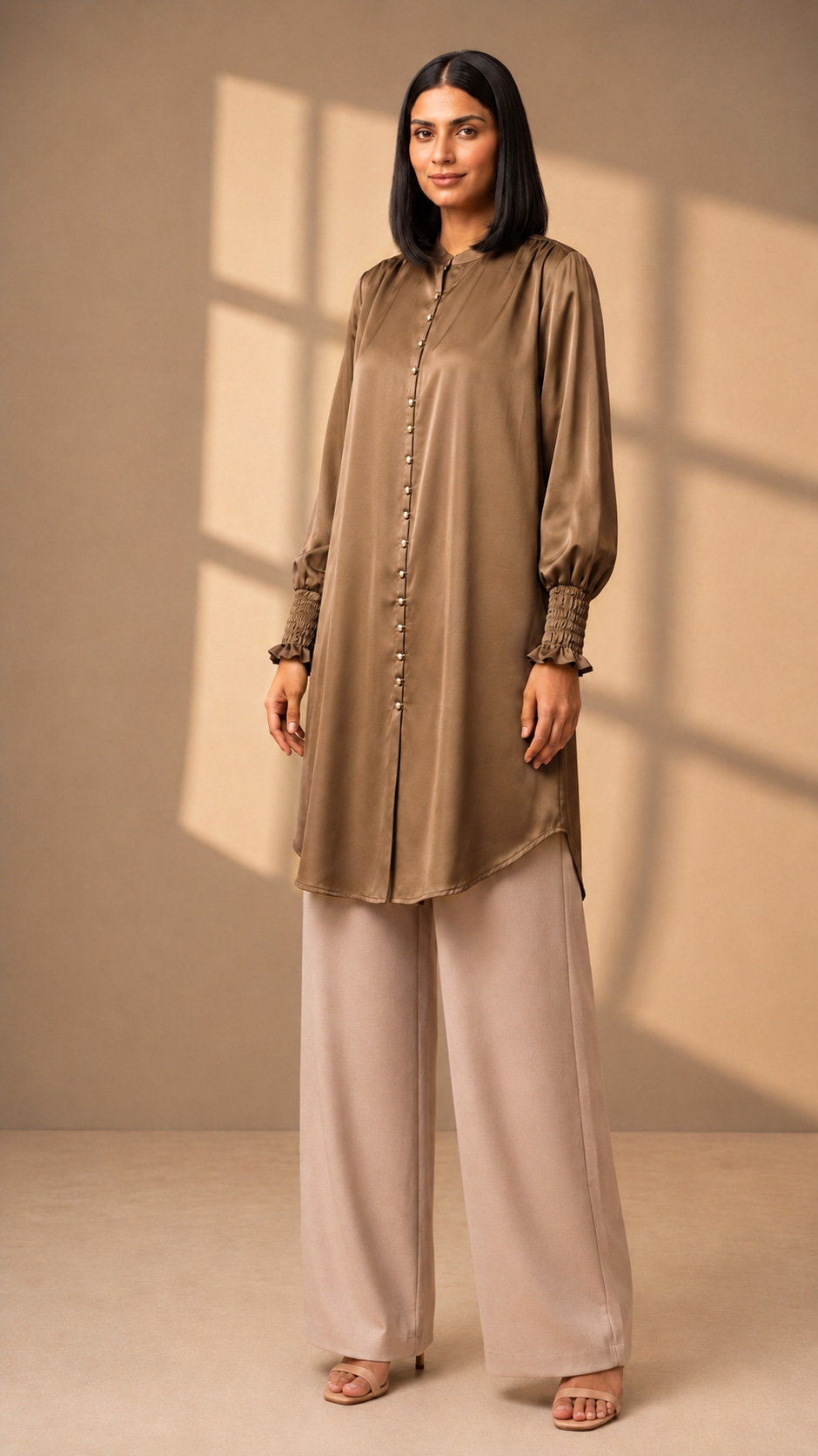 Longline button-down tunic with a mandarin collar, delicate front button placket, long bishop-style sleeves with smocked elastic cuffs and subtle ruffle trim, finished with a gently curved hem. Styled with wide-leg beige trousers.